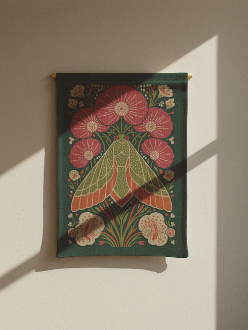 A rectangular woven wall tapestry with a rich, dark green background. The central design is a large, stylized moth with intricate wings in shades of orange, cream, and muted green. The moth is surrounded by a symmetrical arrangement of folk-art style flowers and botanicals in vibrant pink, coral, and cream. The entire piece has a visible, high-quality woven texture. This polyester and cotton blend textile art measures 26x36 inches and includes a rod pocket on the black backing for easy hanging.